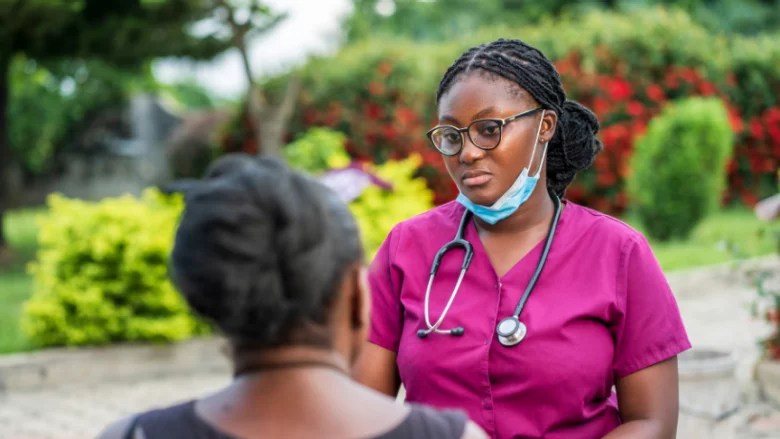 A healthcare professional in a purple scrubs and glasses listens attentively to a patient in a garden setting.