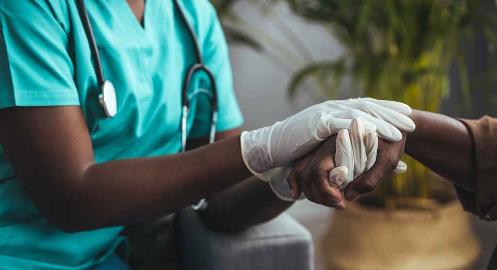 A healthcare professional in scrubs wearing gloves gently holds the hand of a patient in a comforting gesture.